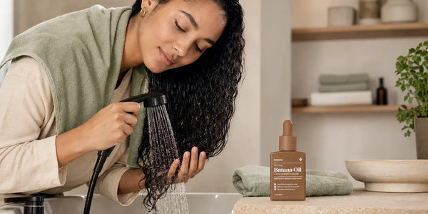 Woman rinsing oil from her hair with lukewarm water beside a bottle of Keyoma Batana Oil.