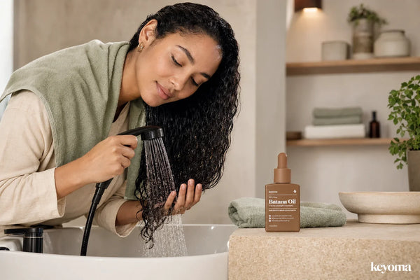 Woman rinsing oil from her hair with lukewarm water beside a bottle of Keyoma Batana Oil.