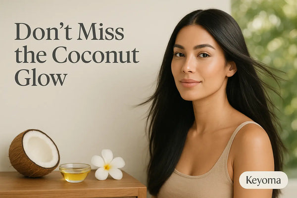 A woman with long glossy hair sits beside a halved coconut, a small dish of oil, and a white flower, illustrating the coconut-oil glow message from Keyoma.