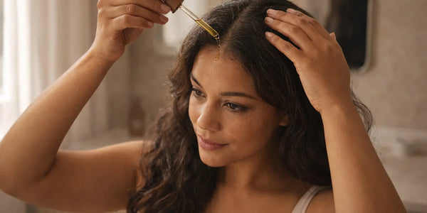 Woman applying oil to her scalp with a dropper in a bathroom setting with soft window light.